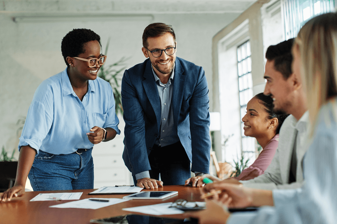 Equipe diversa em reunião de trabalho, com dois colaboradores de pé a liderar a discussão em torno de uma mesa.