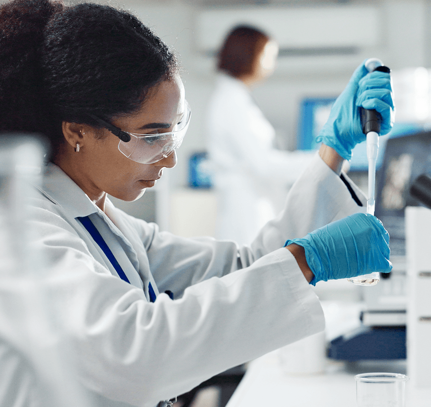 Cientista mulher com bata branca, óculos de proteção e luvas azuis a trabalhar com pipeta em laboratório.