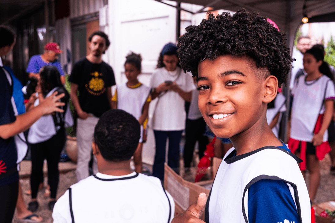 Jovem a sorrir em primeiro plano, rodeado por um grupo de crianças e adolescentes num ambiente comunitário ao ar livre.